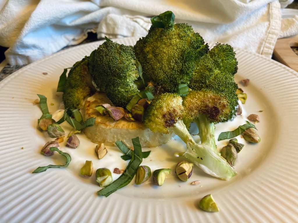 White plate with roasted broccoli and celery root over yogurt with chopped pistachios and basil chiffonade