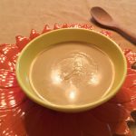 yellow bowl of curried cauliflower soup on an orange flower-shaped charger with a wooden spoon